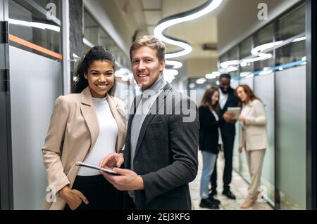 Portrait of two multiracial satisfied successful colleagues dressed in formal wear standing in a modern office, looking and friendly smiling at the camera Stock Photo