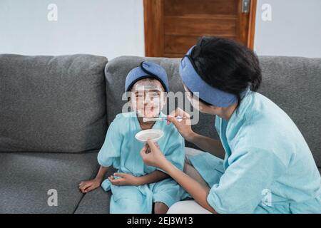 Little girls have fun when a mother smears facial cream with a brush on her face while relaxing together at home Stock Photo