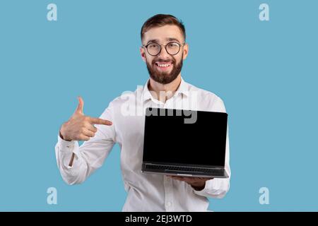Positive young entrepreneur pointing at laptop computer with blank screen for website design on blue studio background Stock Photo