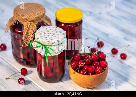 Preserved cherry fruit compote in glass jar on white wooden table, selective focus. Homemade canned organic red cherries in glass jar. Stock Photo