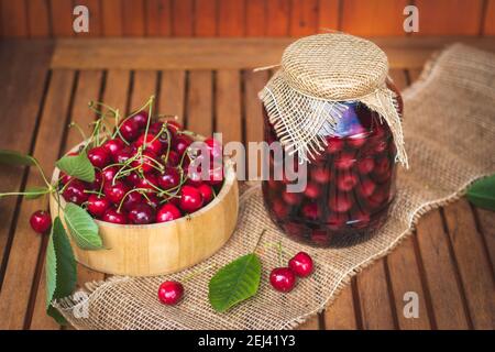 Preserved cherry in jar and fresh harvested red cherries on wooden table. Canned food. Sour fruit compote Stock Photo