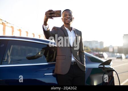 Portrait of young handsome black man in business suit, making selfie photo on his smartphone while leaning on his electric car, charging the battery Stock Photo