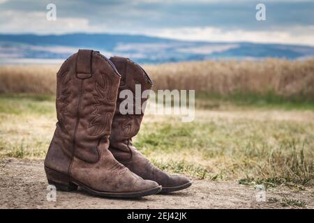 Cowboy boot in rural scene with moody sky. Old brown leather boots outdoors. Wild west retro style Stock Photo