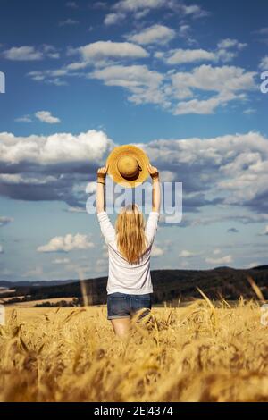 Happy woman holding straw hat is standing in wheat field. Enjoyment of summer. Carefree blond hair woman wearing casual clothing against beautiful sky Stock Photo