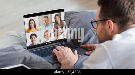 Young man having online work meeting on laptop at home, lying in bed and having group video call with his coworkers Stock Photo