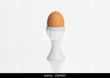 A brown farm egg in a white egg cup isolated on a plain white surface and background. Stock Photo