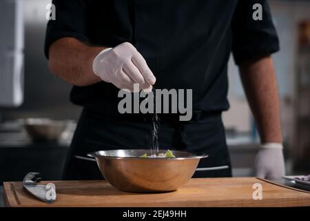 chef cook hands in gloves adding salt in greek salad at restaurant kitchen Stock Photo