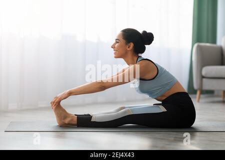 Sporty woman sitting on yoga mat, stretching at home Stock Photo