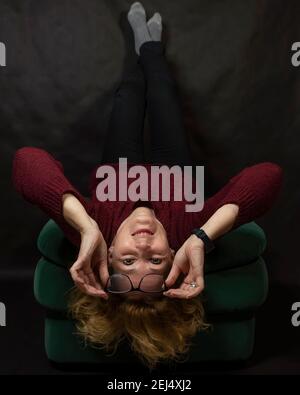 A middle-aged white woman is lying upside down on a pouf while holding her glasses Stock Photo
