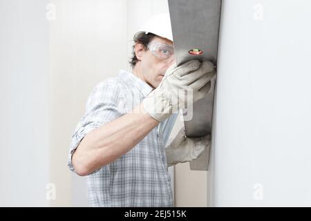 Man drywall worker or plasterer checking level of white plasterboard wall with bubble level at construction site. Wearing white hardhat, work gloves a Stock Photo