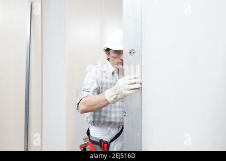 Man drywall worker or plasterer checking level of white plasterboard wall with bubble level at construction site. Wearing white hardhat, work gloves a Stock Photo