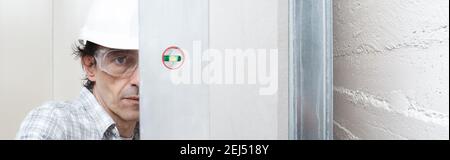 Man drywall worker or plasterer checking level of white plasterboard wall with bubble level at construction site. Wearing white hardhat and safety gla Stock Photo
