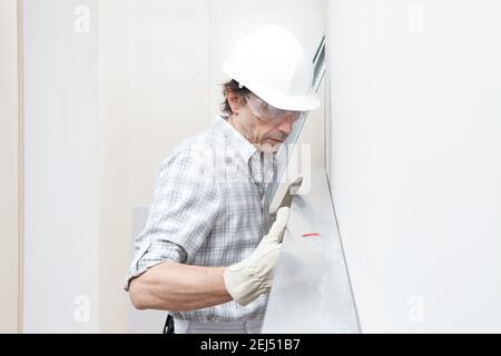 Man drywall worker or plasterer checking level of white plasterboard wall with bubble level at construction site. Wearing white hardhat, work gloves a Stock Photo