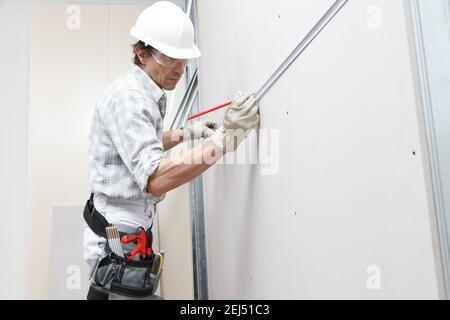 Side view of male drywall worker or plasterer taking measures on plasterboard white wall with meter and pencil at construction site. Wearing white har Stock Photo