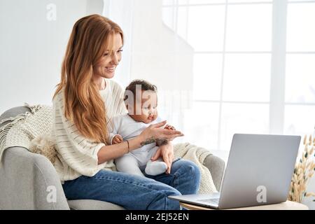 Happy mother holding cute baby daughter looking at laptop computer at home. Stock Photo