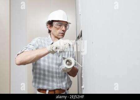 man drywall worker or plasterer putting mesh tape for plasterboard on a wall using a spatula and plaster. Wearing white hardhat, work gloves and safet Stock Photo