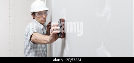 man drywall worker or plasterer sanding and smoothing a plasterboard walls with stucco using a sandpaper holder. Wearing white hardhat and safety glas Stock Photo