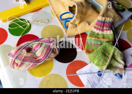 Ball of wool sitting on a table with knitting needles, partly knit sock, craft yarn making warm woolly handmade woollen socks during covid pandemic UK Stock Photo