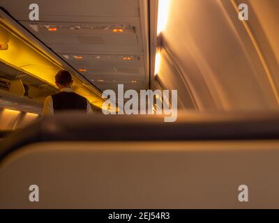 Stewart standing in the aisle of an airplane aircraft cabin. Overhead baggage rack opened and yellow lights turned on. Stock Photo