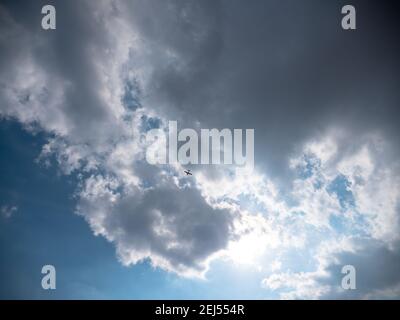 Airplane flying high up in the sky below dark blue stormy clouds with sun shining through them. Stock Photo