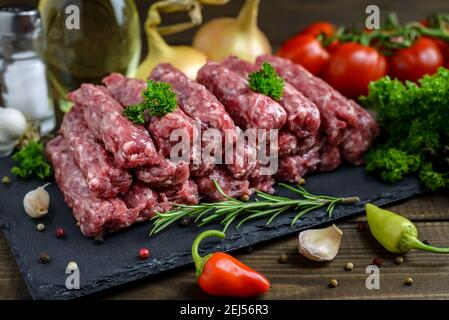 Raw minced meat cevapi ready for barbeque with various fresh vegetables on a wooden table. Selective focus. Stock Photo