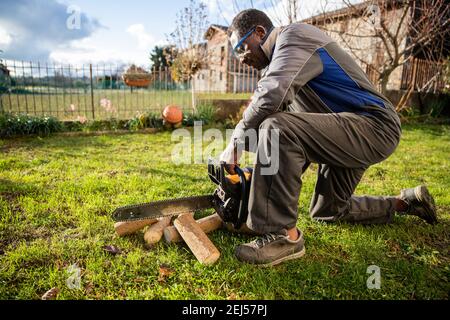 African woodcutter on his knees cuts a piece of wood with his chainsaw Stock Photo