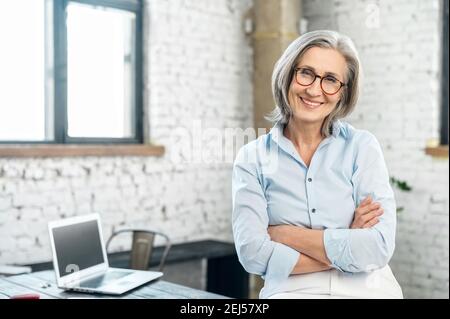 Portrait of a charming smiling elegant lady or mature gray-haired positive businesswoman, standing in the office at her workplace with arms folded Stock Photo