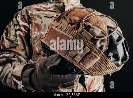 Photo of soldier in camouflaged uniform and tactical gloves holding military helmet with cover on black background. Stock Photo