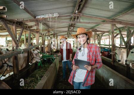 farmer girl standing and smiling with a tablet with the background of the farmer boy feeding grass to the cows in the cow farm shed Stock Photo