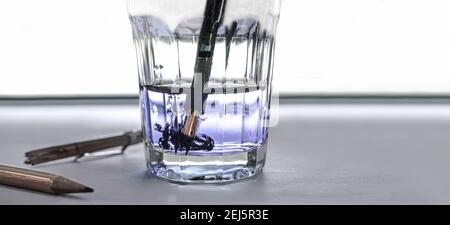 on a surface we have a broken pencil, a glass of water in which the nib of the pen has been plunged. The ink mixes quietly with the water in the glass Stock Photo