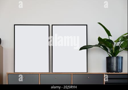 Two vertical black mockup frames on wooden design sideboard leaning against white wall next to green plant in Scandi design Stock Photo