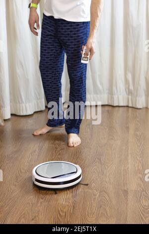 Man does house cleaning, turns on cordless robot vacuum cleaner using remote control. Concept of everyday cleaning at home, helping men around house, Stock Photo