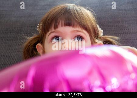 A cute, brown-haired, blue-eyed baby girl sitting on a sofe and hading her face behind a big pink balloon. Stock Photo