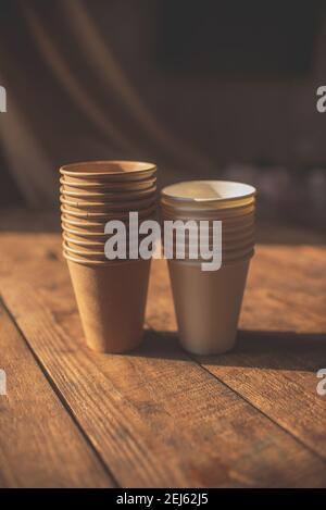 disposable cups made of dark brown paper and white cups stand on wooden table against brown background Stock Photo