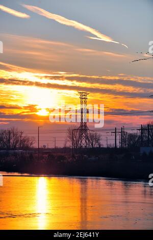 Bartlett, Illinois, USA. The sun setting behind a large power line tower and reflecting into a pond in an industrial park. Stock Photo