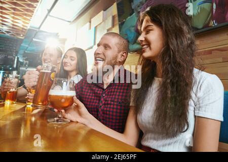 Side view of cheerful friends laughing and joking while drinking ale in pub. Happy men and woman sitting together in bar and toasting on weekends. Concept of happiness and fun. Stock Photo