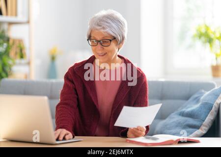 Elderly woman, sitting on the sofa with a paper receipt in her hands, are calculating expenses, managing the family budget. Stock Photo