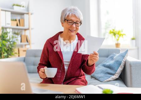 Elderly woman, sitting on the sofa with a paper receipt in her hands, are calculating expenses, managing the family budget. Stock Photo
