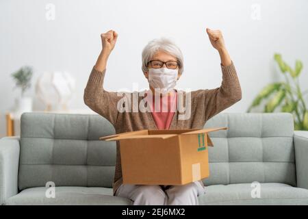 Senior woman is holding cardboard box sitting on sofa at home. Stock Photo