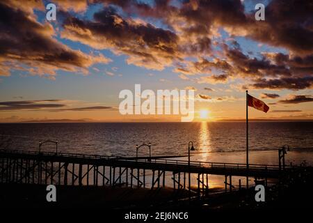 Walkers on White Rock's iconic pier enjoy stunning sunset over the sea.  In the foreground, Canada's national flag proudly flutters in the breeze. Stock Photo