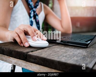 Finger using modern white wireless mouse while working on black laptop computer on wooden desk. Female’s hand clicking on mouse. Stock Photo