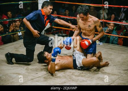 Muay Thai fight in isan, rural Thailand, Thai boxing, the referee trying to stop boxer from punching opponent lying on the ground. Stock Photo