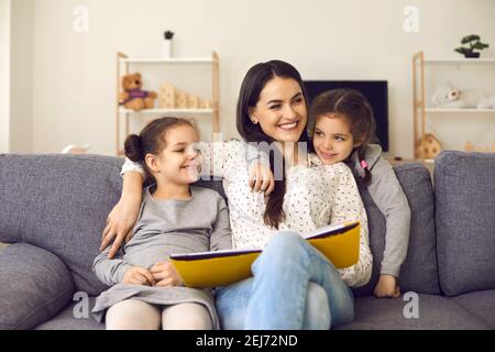 Smiling cheerful mother and small daughters sitting on sofa with book, hugging and looking aside Stock Photo