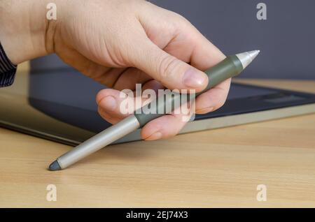 A man's hand with a special pen for a graphics tablet in close-up. A professional device for designers lies in the background. Side view from an angle Stock Photo