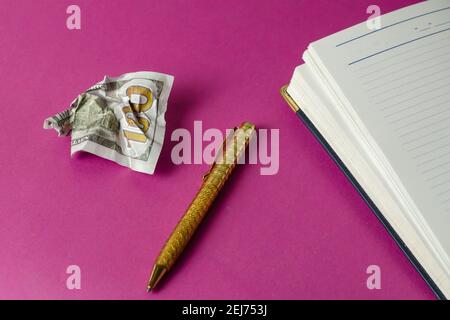 Open diary, gold pen and crumpled one hundred US dollar bill on pink background. Top view at an angle. Selective focus Stock Photo