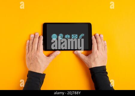 Male hand holding a black digital tablet displaying contact and communication icons on yellow background. Stock Photo