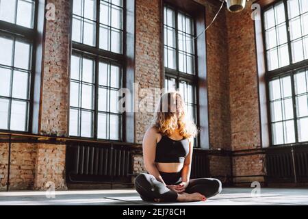 Young curly female sit in lotus pose on floor of loft hall and smile. Positive, meditation, concentrate Stock Photo
