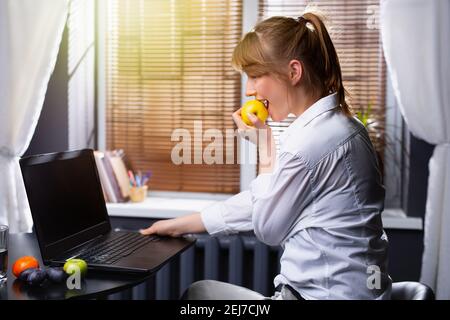 The blonde girl works at home while sitting at a laptop and eats an apple. Snack while working online. Remote work during quarantine. Stock Photo