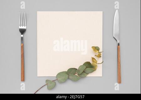 Knife and fork with white paper for menu or recipe text and gold eucalyptus branch on neutral grey table. Mockup Blank card and cutlery. Stock Photo