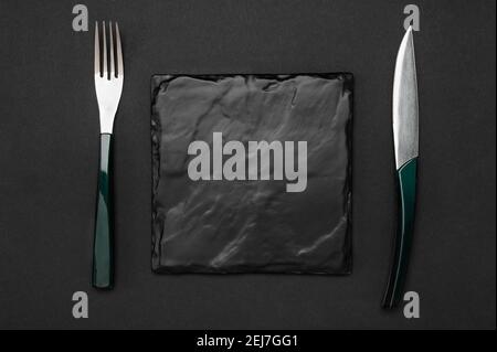 Knife and fork with square black slate board for menu or recipe text on black table. Mockup Blank board and cutlery on black table background Stock Photo